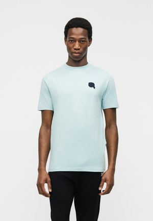 Junger Mann, der ein hellblaues Kurzarm-T-Shirt mit kleinem dunkelblauem Logo auf der Brust und schwarze Hose trägt, vor einem einfarbigen Hintergrund stehend.