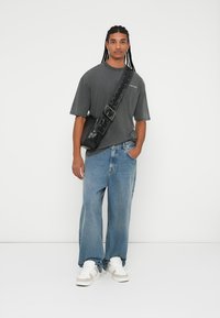 Sivý oversized tričko, široké svetlomodré džínsy, čierna crossbody taška so strapcom a biele tenisky, na pozadí bez vzoru.