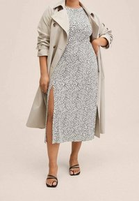 Trench-coat beige porté sur une robe blanche à motif moucheté noir, avec une fente haute jusqu'à la cuisse. Sandales noires à lanières aux pieds.