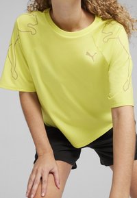 Person klädd i en ljusgul Puma-t-shirt med kattlogotyper på ärmarna och bröstet, ihop med svarta shorts, lutad framåt.