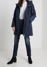 Abrigo acolchado azul marino con capucha, cierre de cremallera y dos bolsillos delanteros. Lleva puesto sobre una camisa blanca y combinado con jeans oscuros y botas de tobillo.