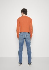 Oranžová mikina s kulatým výstřihem, doplněná modrými džínami s kapsami na zadní straně. Oblečení je doplněno černo-bílými teniskami.