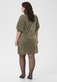 Olijfgroen corduroy mini-jurk met verticale strepen, pofmouwen en een losse fit. Draagt met sheer zwarte panty’s en zwarte sleehakken.