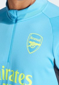 Ljusblå sporttröja med dragkedja vid kragen, gul Arsenal-logga och texten "Emirates" i starkt gult över bröstet.
