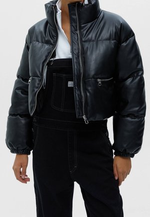 Persoon die een zwarte ingekorte donsjas met ritszakken draagt over een zwarte overall en een wit shirt met ritssluiting, staand tegen een effen achtergrond.