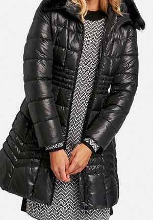 Vrouw draagt een zwart doorgestikt donzen jas met bontkraag over een zwart-witte jurk met zigzagpatroon, handen zachtjes samengevouwen voor zich.