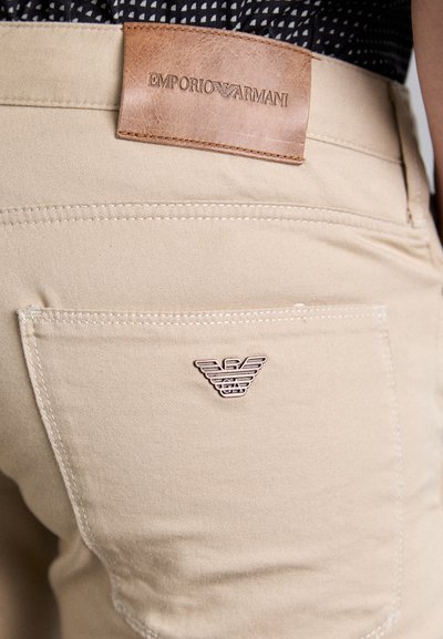 Bež pamuk hlače imaju kožnu oznaku Emporio Armani na pojasu i mali metalni logotip na stražnjem džepu.