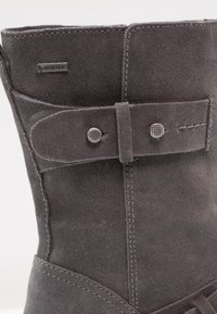 Botte en daim gris avec étiquette Gore-Tex, dotée d'une sangle décorative ornée de deux clous en métal texturé près du sommet.