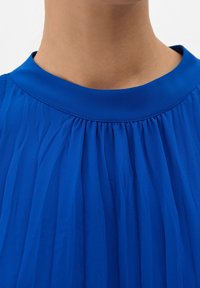 Blusa blu con collo alto e rotondo, in tessuto plissettato, che presenta una texture liscia e un colore vivace. Nessun adornamento visibile.