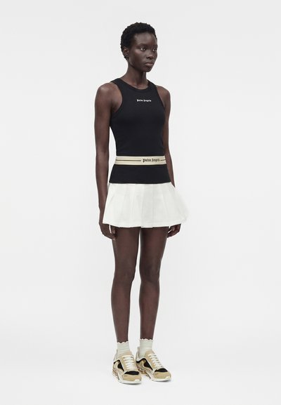 Γυναίκα όρθια, φορά μαύρο αμάνικο τοπ και λευκή πλισέ φούστα με διαφημιστική ζώνη, σετ με μπεζ, μαύρα και λευκά αθλητικά παπούτσια και ανοιχτές κάλτσες.