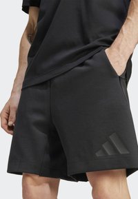 Melnas sporta šortas ar mīkstu faktūru, kas aprīkoti ar sānu kabatu un spīdīgu melnu trīs svītru logotipu apakšmalā.