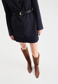 Brązowe zamszowe botki do kolan z szpiczastymi noskami. Noszone z ciemną mini spódnicą i żakietem, podkreślone czarnym paskiem. Gładka tekstura.