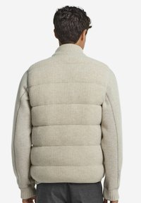 Beige gestrickte Daunenjacke mit Rippkragen und seitlichen Einsätzen; strukturiertes, gepolstertes Design mit engem Schnitt und langen Ärmeln.