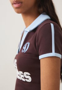 Vrouw draagt een bruin poloshirt met lichtblauwe kraag en mouwzoom, met gedeeltelijke tekst en nummer "97" op de borst.