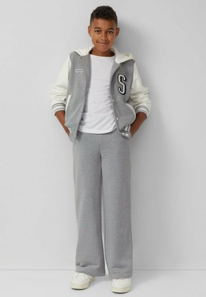Grijze varsityjack met witte mouwen, met een "S" op de borst, gecombineerd met grijze joggingbroek. Witte sneakers, een eenvoudig wit T-shirt eronder.