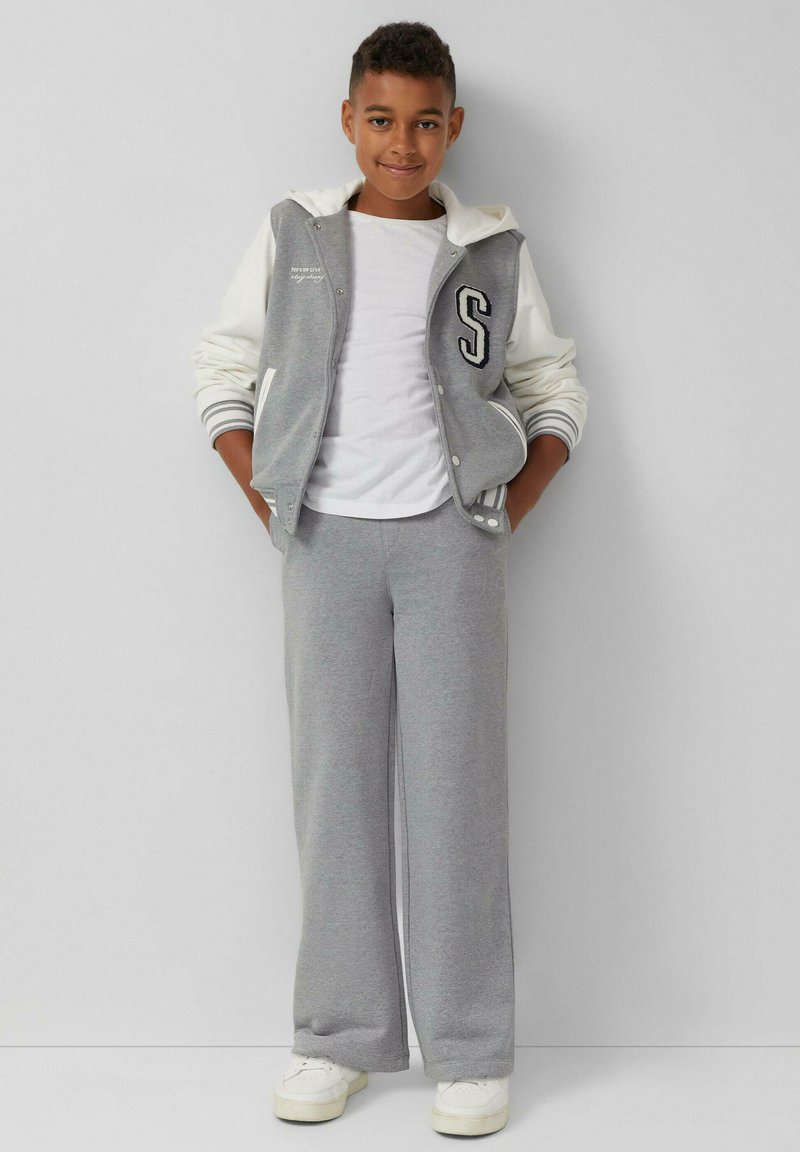 Grijze varsityjack met witte mouwen, met een "S" op de borst, gecombineerd met grijze joggingbroek. Witte sneakers, een eenvoudig wit T-shirt eronder.