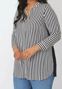 Blusa a righe bianco e nero con scollatura a V, maniche lunghe e spacchetti laterali. Il tessuto ha una texture liscia, abbinata a jeans in denim blu.
