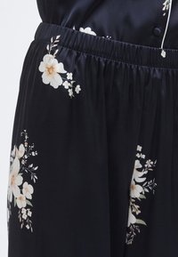 Tissu noir à imprimé floral, avec des fleurs blanches et beige. Taille élastique, texture lisse et coupe décontractée.