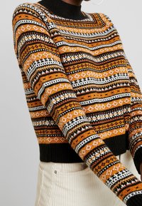 Neulepaita, jossa on korkea musta kaulus, oranssia, mustaa ja valkoista geometrista kuviota, sekä resorit hihansuissa ja helman päällä.