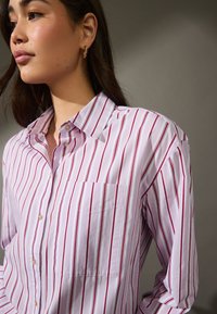 Camicia a righe con base rosa chiaro e strisce verticali rosse. Presenta un colletto a punta e una tasca sul petto. Materiale in cotone.