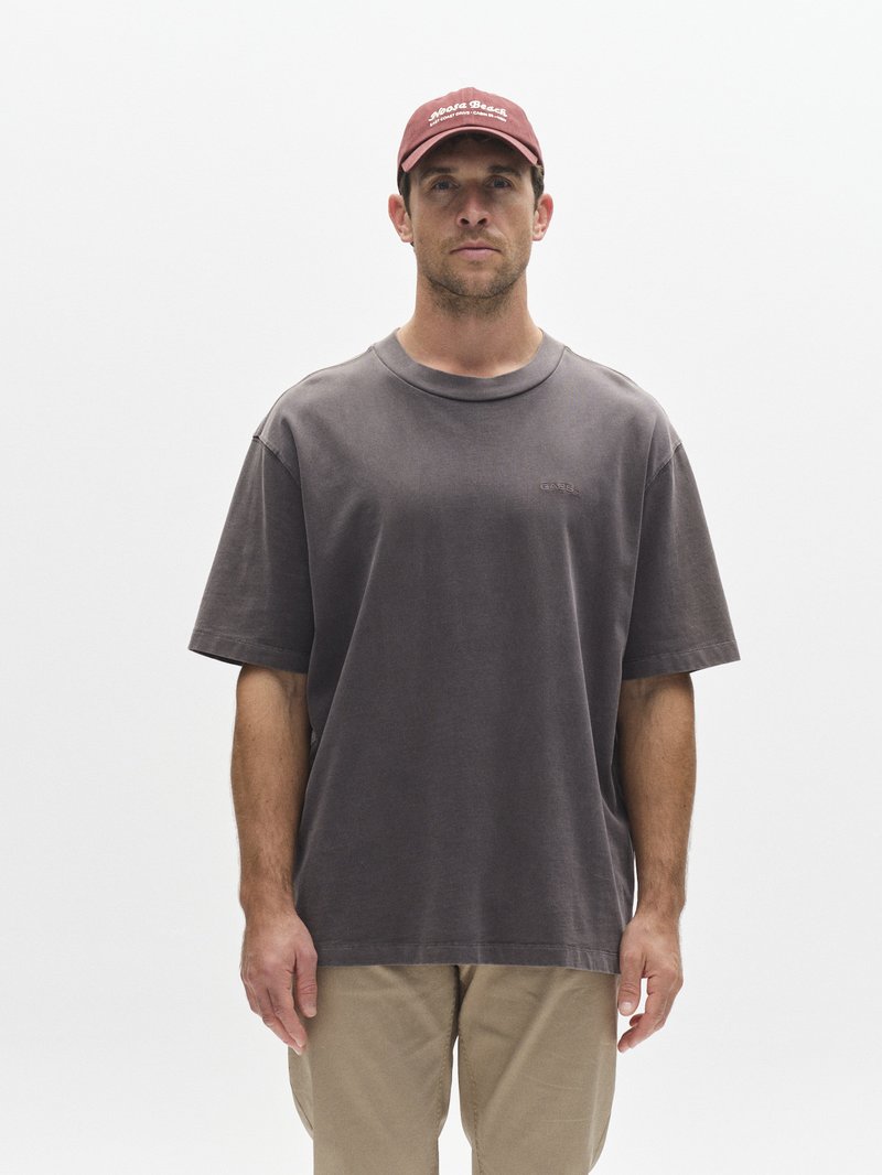 Oversized grijs T-shirt, katoenen materiaal, ronde hals, korte mouwen. Gecombineerd met lichtgekleurde broek en een kastanjebruine pet met witte tekst.