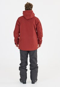 Rote wasserdichte Jacke mit Kapuze, lockere Passform und langen Ärmeln, kombiniert mit grauen Hosen. Glatte Textur, praktisches Design.