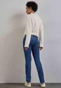 Kermanauha kuviollinen neule, siniset farkut takataskuilla ja vaaleat lenkkarit. Kankaan rakenne näyttää pehmeältä ja rentoilta designilta.