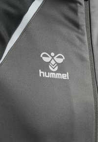 Giacca atletica grigia con logo bianco, texture liscia, frontale con zip e dettagli in grigio chiaro a contrasto lungo le spalle.