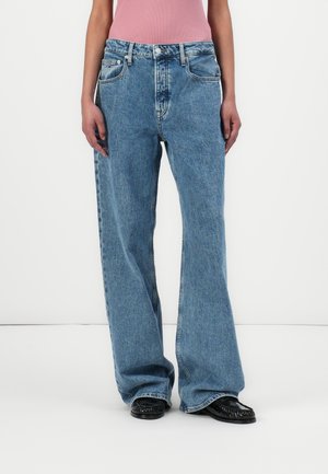 Persona que lleva vaqueros azules holgados, zapatos negros y una camiseta sin mangas rosa, de pie sobre un fondo blanco liso.