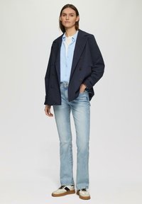 Marineblauwe double-breasted jas, lichtblauw button-up shirt, vervaagde blauwe flared jeans, en beige sneakers met zwarte Accenten. Gladde texturen.