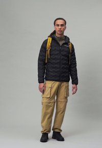 Black puffer jacket with quilted pattern, tan cargo pants, and a yellow backpack. Black footwear completes the outdoor outfit.