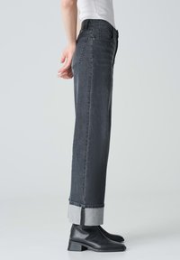 Jeans grigio scuro a vita alta con taglio dritto, caratterizzati da risvolti che mostrano un tessuto più chiaro. Indossati con stivaletti neri alla caviglia.