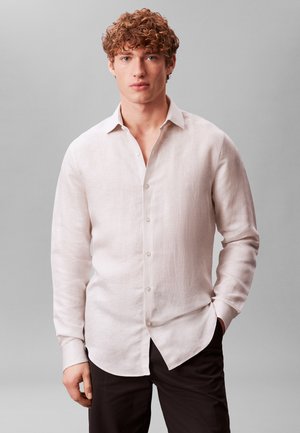 Νέος άνδρας με κατσαρά μαλλιά, φορώντας ένα ανοιχτό μπεζ πουκάμισο με κουμπιά και μαύρο παντελόνι, στέκεται μπροστά σε μονόχρωμο γκρι φόντο.