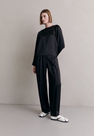 Haut en satin noir à manches longues avec un col rond et une coupe décontractée, assorti à un pantalon large noir ; modèle portant des sandales blanches avec des accents noirs.