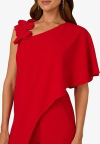 Červený kombinéza s asymetrickým designem, který má volnou plášťovou rukáv a látkové květiny na jednom rameni. Hladká textura, živá barva.
