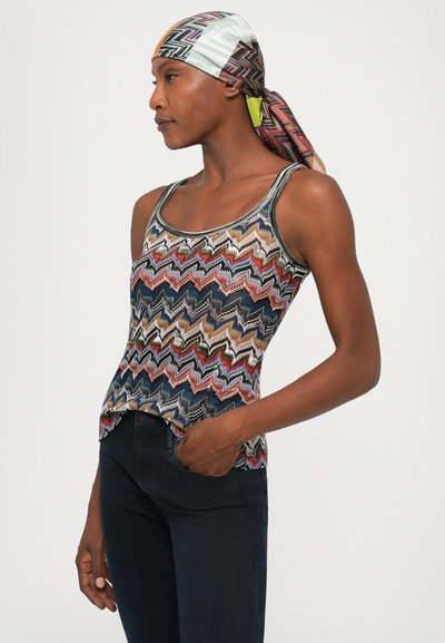 Kvinna som bär en färgglad väst med zigzag-mönster, mörka jeans och en flerfärgad geometrisk mönstrad huvudduk, stående med handen i fickan.