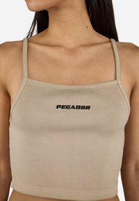 Beige ribbed crop top with slim straps and a black "PEGADOR" logo centered on the front. Smooth texture and fitted design.