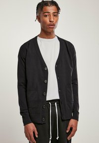 Fekete kardigán puha anyagból, V-nyakkal, hat gombos záródással és két elülső zsebbel. Fehér ing és fekete nadrág fölött viselve.