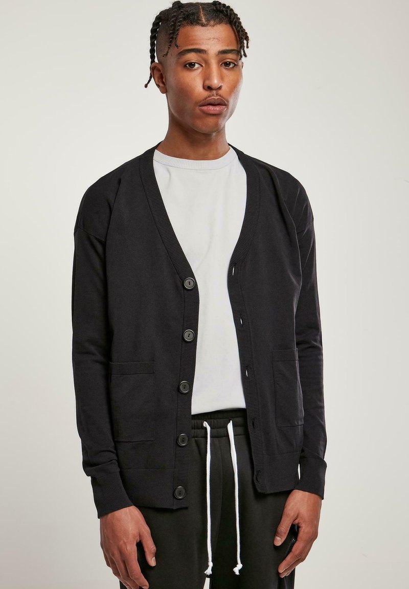 Fekete kardigán puha anyagból, V-nyakkal, hat gombos záródással és két elülső zsebbel. Fehér ing és fekete nadrág fölött viselve.