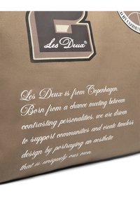 Barna vászontáska fehér szöveggel. Nagy logótervezéssel és márkázott hímezéssel rendelkezik. Sima tapintású, tartós anyag.