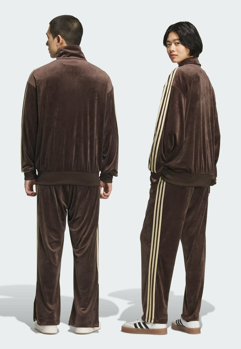 Bruine velours trainingspak met hoge kraag, voorzien van crèmekleurige strepen langs de mouwen en de broek. Losse pasvorm en textuurdetail in de stof.