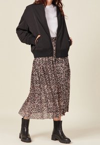 Giacca bomber nera con polsini a costine, abbinata a una gonna maxi a pieghe con stampa leopardata in tonalità terrose, e stivaletti neri con suola spessa.