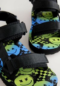 Svarta remsandaler med ett mönstrat fotbädd som har blåa och gröna smileys och ett rutigt mönster, tillverkade av syntetmaterial.