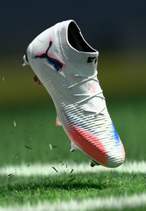 Scarpino da calcio bianco con gradiente rosso e blu, logo Puma nero e colletto alla caviglia simile a una calza, che fluttua sopra l'erba con l'erbetta che si solleva.