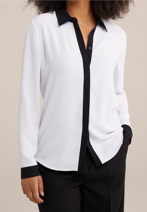 Femme portant un chemisier blanc à manches longues avec col noir, patte de boutonnage noire et poignets noirs, associé à un pantalon noir, main dans la poche.