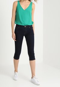 Turquoise mouwloos topje met een V-hals, gecombineerd met donker denim capribroek. Model draagt witte sneakers; gladde stof en een aansluitend ontwerp.