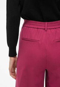 Pantalon magenta avec une ceinture élastique, doté de deux poches arrière et d'une texture lisse. Le tissu semble léger et confortable.