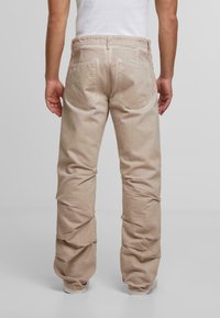 Mies, joka pukeutuu vaaleanbeigeihin suoriin housuihin ja valkoisiin lenkkareihin, seisoo selkä kameraan päin vaaleaa taustaa vasten.