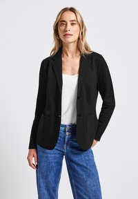 Černý blazer s přizpůsobeným designem, dvěma předními kapsami a dvěma knoflíky. Hladká látka a strukturovaný silueta v kombinaci s lehkými džíny.