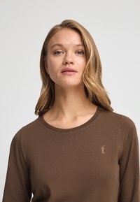 Brun långärmad skjorta med rund halsringning och en liten, gyllene logotyp på vänster bröst. Tyget ser slätt och lätt ut.